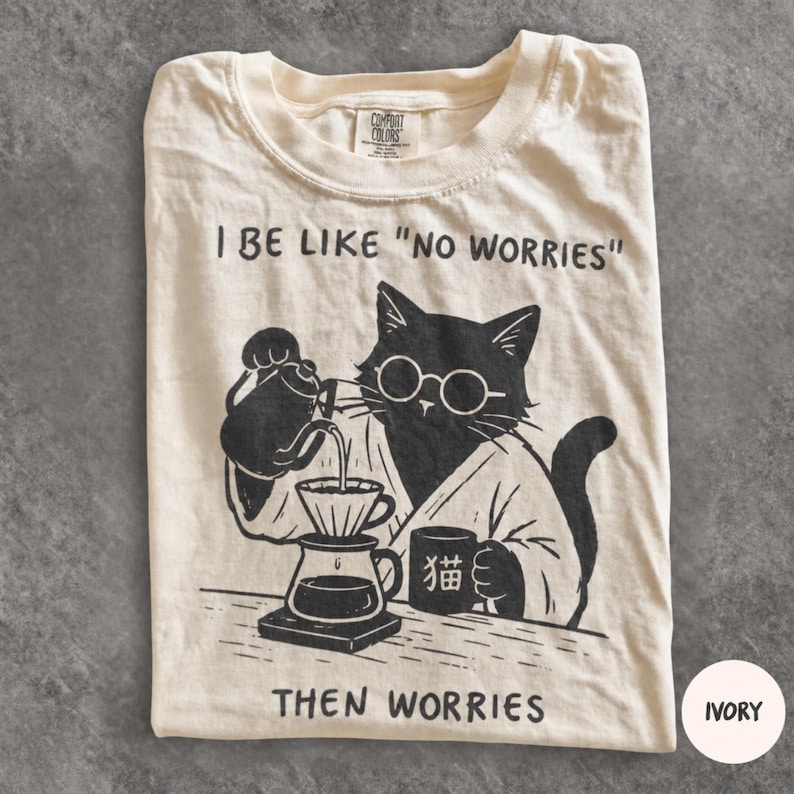 May include: An ivory t-shirt featuring a black graphic of a cat wearing glasses and a robe, pouring coffee into a dripper. The text reads I BE LIKE "NO WORRIES" above the cat and THEN WORRIES below. The cat holds a mug with the Japanese character for cat.
