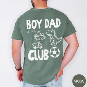 May include: Moss green t-shirt with the words "BOY DAD CLUB" in white, along with a dinosaur, soccer ball, toy truck, and building blocks graphic. The text "est. 2026" is also included.