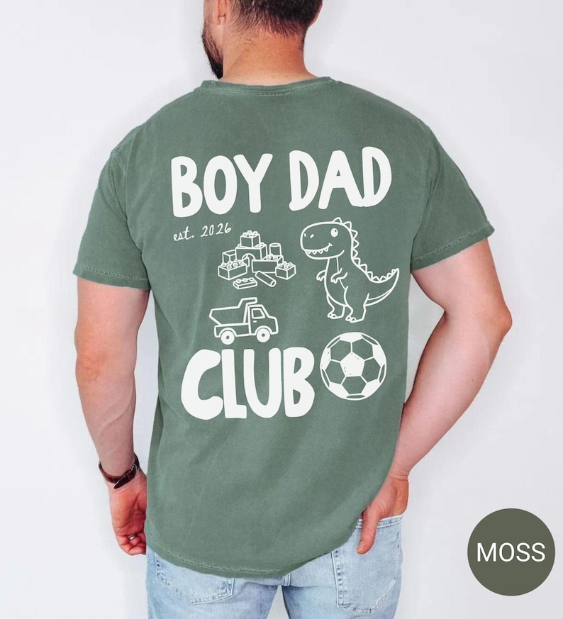 May include: Moss green t-shirt with the words "BOY DAD CLUB" in white, along with a dinosaur, soccer ball, toy truck, and building blocks graphic. The text "est. 2026" is also included.