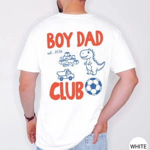 May include: White t-shirt with the words "BOY DAD CLUB" in red and blue. The shirt features a dinosaur, toy blocks, a truck, and a soccer ball graphic. The text "est. 2026" is also included.
