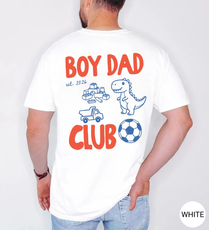 May include: White t-shirt with the words "BOY DAD CLUB" in red and blue. The shirt features a dinosaur, toy blocks, a truck, and a soccer ball graphic. The text "est. 2026" is also included.