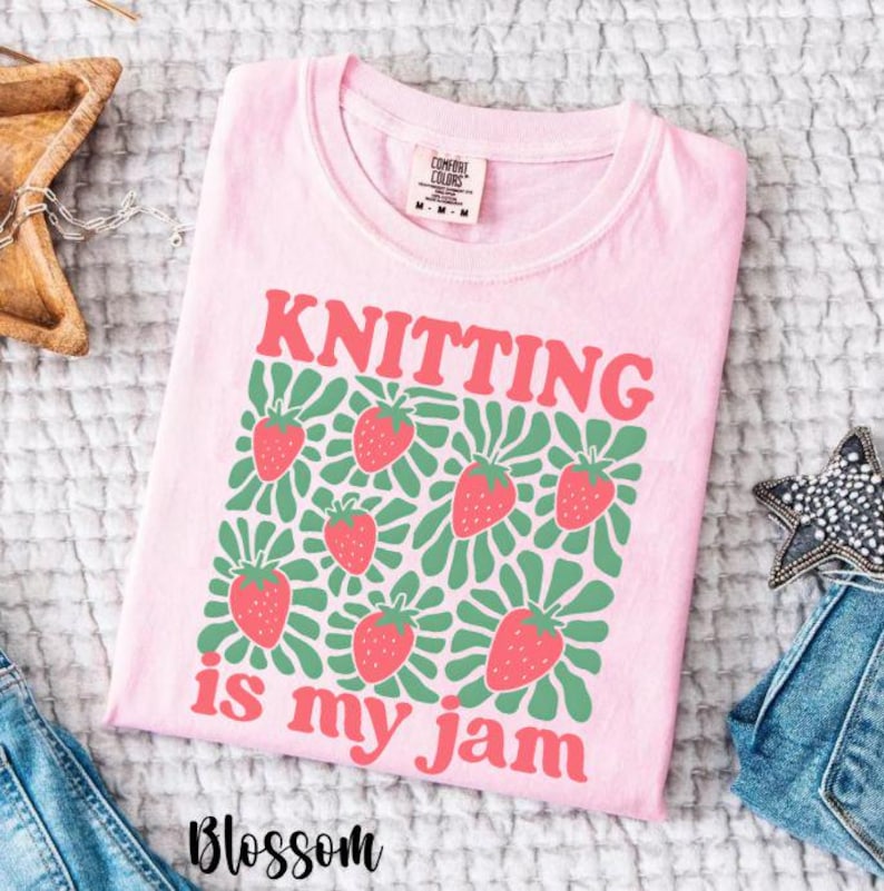 May include: A light pink t-shirt with the words "KNITTING is my jam" in red and green, along with a pattern of strawberries. The shirt is made of a soft material and is a Comfort Colors brand.