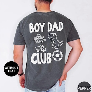 May include: Dark gray t-shirt with the words "BOY DAD CLUB" in white. The shirt features white illustrations of building blocks, a dinosaur, a truck, and a soccer ball. The shirt is made of a soft material.