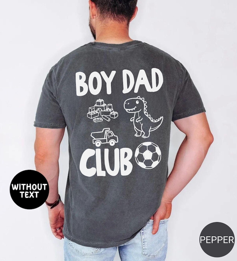 May include: Dark gray t-shirt with the words "BOY DAD CLUB" in white. The shirt features white illustrations of building blocks, a dinosaur, a truck, and a soccer ball. The shirt is made of a soft material.