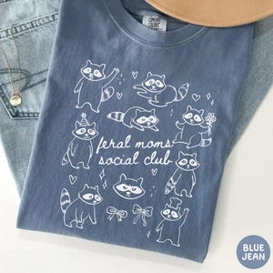 K&ouml;nnte beinhalten: Blaues Jeans-T-Shirt mit einer wei&szlig;en Grafik von Waschb&auml;ren und dem Text "feral moms social club". Das Shirt hat einen Rundhalsausschnitt und kurze &Auml;rmel. Das Shirt besteht aus einem weichen, bequemen Material.