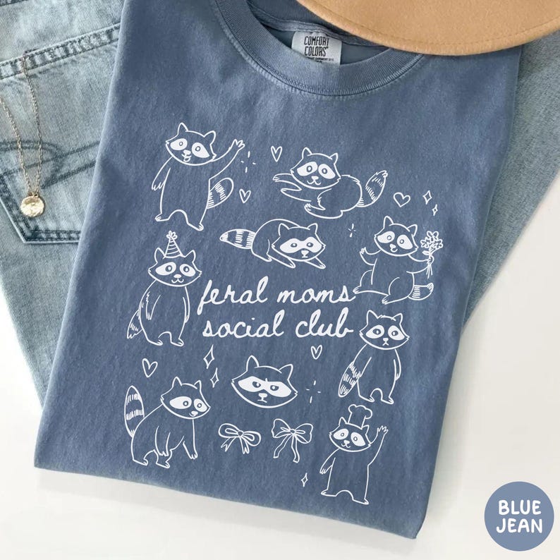 K&ouml;nnte beinhalten: Blaues Jeans-T-Shirt mit einer wei&szlig;en Grafik von Waschb&auml;ren und dem Text "feral moms social club". Das Shirt hat einen Rundhalsausschnitt und kurze &Auml;rmel. Das Shirt besteht aus einem weichen, bequemen Material.