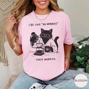 May include: A light pink t-shirt featuring a black graphic of a cat wearing round glasses and a robe, pouring coffee from a kettle into a dripper. The text reads I BE LIKE "NO WORRIES" and THEN WORRIES, with a small mug labeled with Japanese characters.