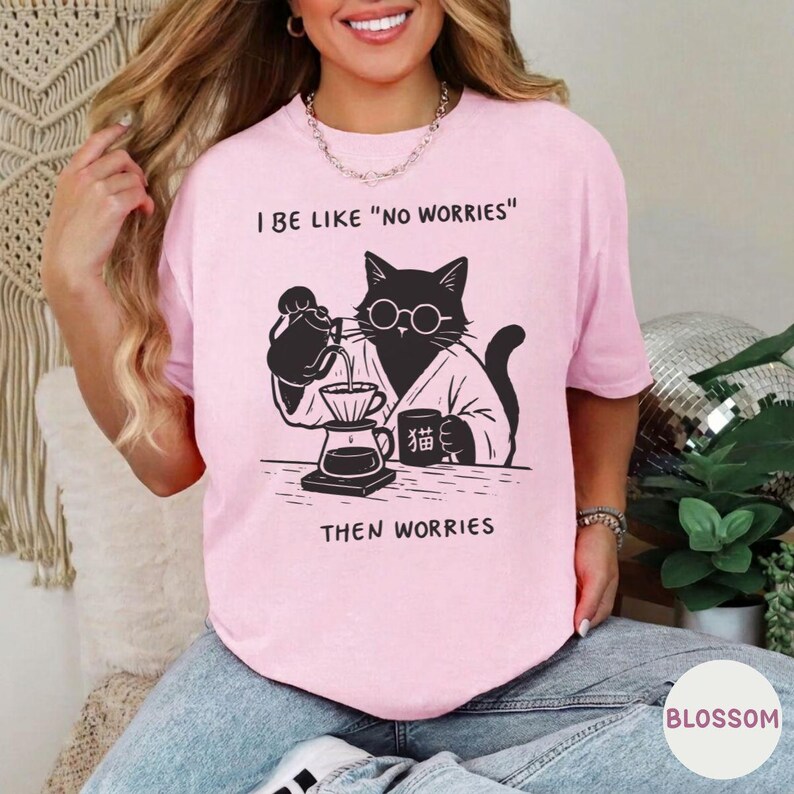 May include: A light pink t-shirt featuring a black graphic of a cat wearing round glasses and a robe, pouring coffee from a kettle into a dripper. The text reads I BE LIKE "NO WORRIES" and THEN WORRIES, with a small mug labeled with Japanese characters.