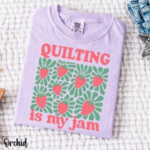 May include: A light purple t-shirt with the text "QUILTING is my jam" in coral. The design includes coral strawberries and green leaves. The shirt is a Comfort Colors brand.