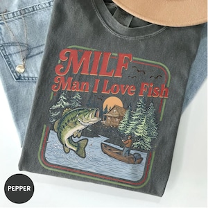May include: Dark gray t-shirt with the text "MILF Man I Love Fish" in red and orange. The graphic includes a fish, a fisherman in a boat, trees, and a cabin. The shirt is folded on top of blue jeans.