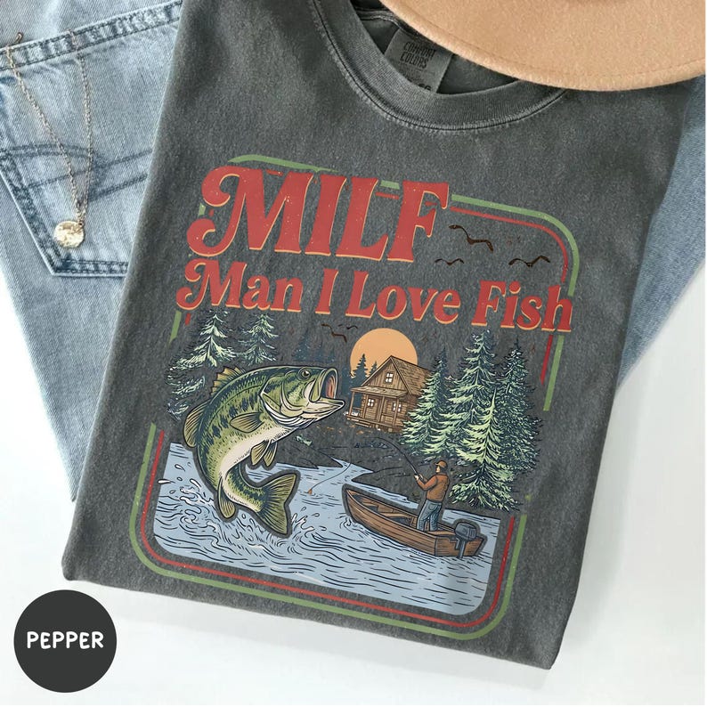 May include: Dark gray t-shirt with the text "MILF Man I Love Fish" in red and orange. The graphic includes a fish, a fisherman in a boat, trees, and a cabin. The shirt is folded on top of blue jeans.