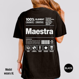 May include: A black t-shirt featuring a white graphic on the back with the text Maestra, 100% Element, Paciente, Creativa, and a list of ingredients including 100% corazon, spiced with humor, and icons representing a book, globe, torch, star, and coffee cup.