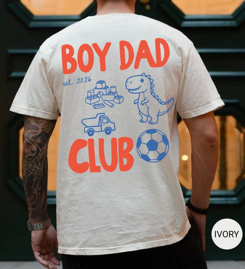 May include: Ivory-colored t-shirt with the words "BOY DAD CLUB" in orange. The shirt features blue illustrations of a dinosaur, toy blocks, a truck, and a soccer ball. The text "est. 2026" is also printed on the shirt.
