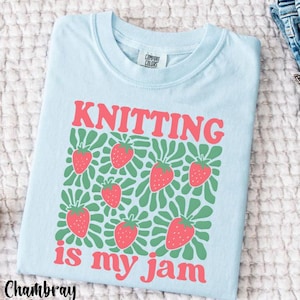 May include: Light blue t-shirt with the words "KNITTING is my jam" in red. The shirt features a pattern of red strawberries with green leaves. The shirt is made by Comfort Colors.