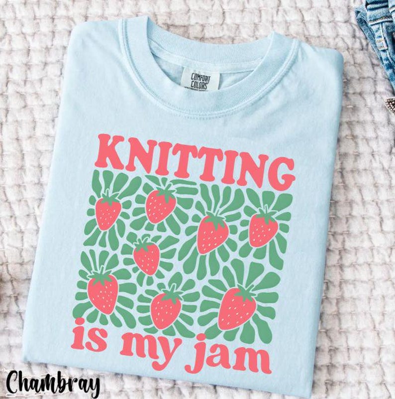 May include: Light blue t-shirt with the words "KNITTING is my jam" in red. The shirt features a pattern of red strawberries with green leaves. The shirt is made by Comfort Colors.