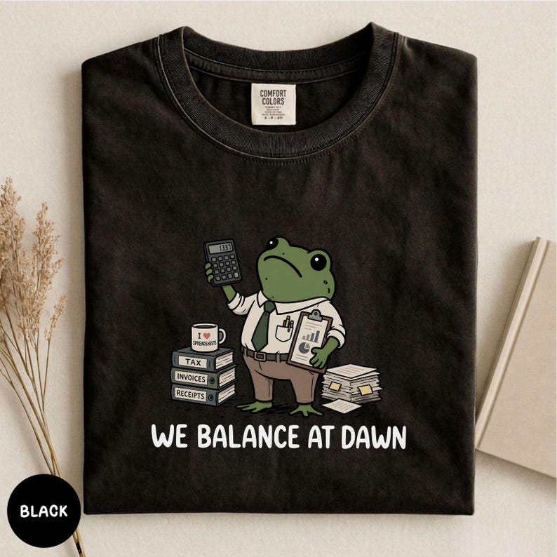 K&ouml;nnte beinhalten: Ein schwarzes T-Shirt mit einer Grafik eines gr&uuml;nen Frosches in wei&szlig;em Hemd und brauner Hose. Der Frosch h&auml;lt einen Taschenrechner und ein Klemmbrett neben B&uuml;cherstapeln mit der Aufschrift Tax, Invoices, Receipts und dem Text WE BALANCE AT DAWN.