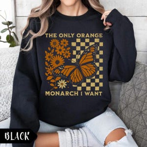 May include: Black sweatshirt with a graphic design featuring a monarch butterfly, flowers, and the text "THE ONLY ORANGE MONARCH I WANT." The design includes orange and yellow elements on a checkered background.