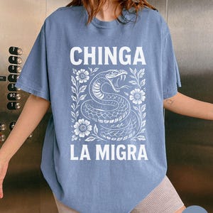 Puede incluir: Una camiseta azul vaquera con las palabras "CHINGA LA MIGRA" en letras blancas. Una ilustraci&oacute;n blanca de una serpiente con flores y hojas est&aacute; centrada debajo del texto. La camiseta tiene mangas cortas.