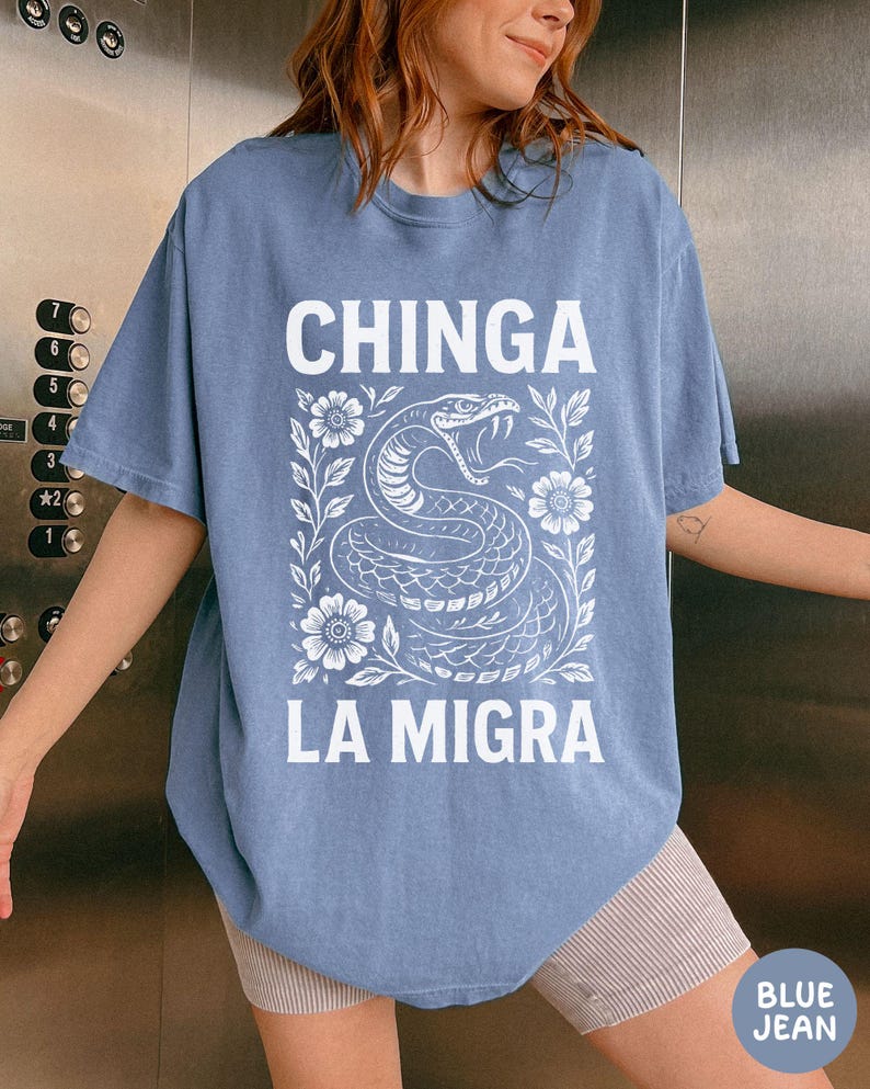 Puede incluir: Una camiseta azul vaquera con las palabras "CHINGA LA MIGRA" en letras blancas. Una ilustraci&oacute;n blanca de una serpiente con flores y hojas est&aacute; centrada debajo del texto. La camiseta tiene mangas cortas.
