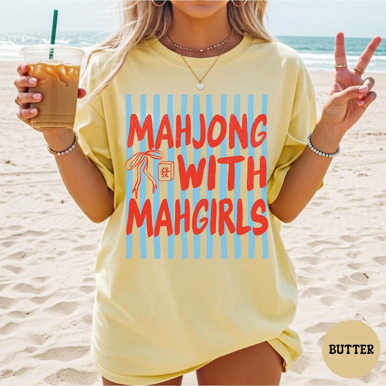 Peut inclure: Un t-shirt &agrave; manches courtes jaune p&acirc;le avec un imprim&eacute; graphique compos&eacute; de rayures verticales bleu clair et du texte rouge "MAHJONG WITH MAHGIRLS", accompagn&eacute; d'une petite illustration de ruban rouge et d'une tuile de mah-jong.