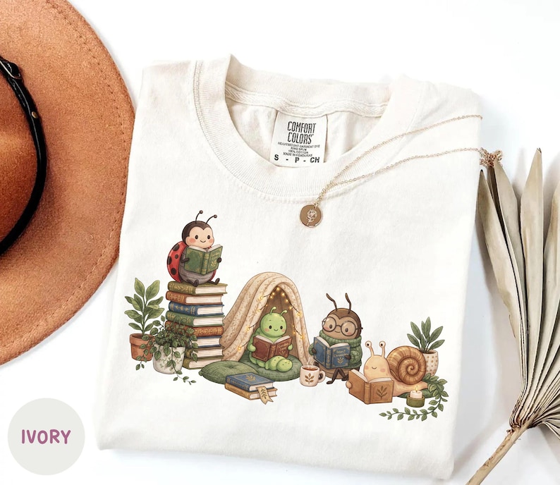 May include: An ivory t-shirt featuring a graphic illustration of a ladybug, a caterpillar in a tent, a beetle with glasses, and a snail, all reading books. The shirt includes a Comfort Colors brand label at the neck and a gold-toned pendant necklace.