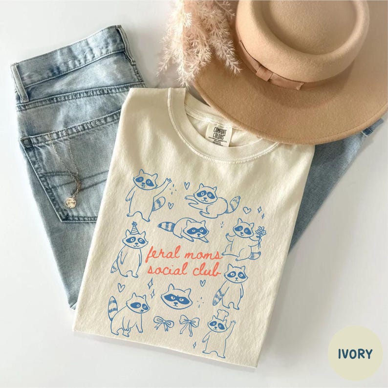 K&ouml;nnte beinhalten: Cremefarbenes T-Shirt mit blauem Waschb&auml;r-Motiv und dem Text "feral moms social club" in Rot und Blau. Das Shirt wird mit hellblauen Jeans und einem beigen Hut kombiniert. Eine silberne Halskette ist sichtbar.