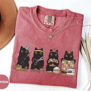 May include: A crimson short-sleeve t-shirt featuring a graphic of four black cats engaged in sewing activities, including holding a needle, scissors, fabric scraps, and a quilt. The inner neck label reads Comfort Colors, 100% Cotton, Made in Honduras.