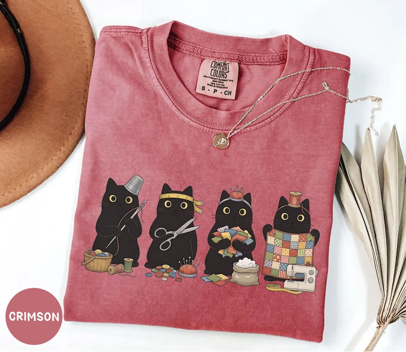 May include: A crimson short-sleeve t-shirt featuring a graphic of four black cats engaged in sewing activities, including holding a needle, scissors, fabric scraps, and a quilt. The inner neck label reads Comfort Colors, 100% Cotton, Made in Honduras.