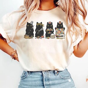 May include: An ivory t-shirt featuring a graphic print of four black cats engaged in sewing activities. The cats are depicted with sewing tools including a thimble, scissors, a pincushion, fabric scraps, a sewing machine, and a colorful quilt.