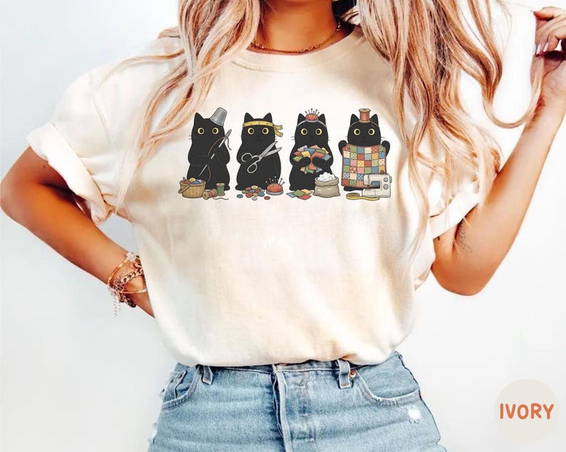 May include: An ivory t-shirt featuring a graphic print of four black cats engaged in sewing activities. The cats are depicted with sewing tools including a thimble, scissors, a pincushion, fabric scraps, a sewing machine, and a colorful quilt.
