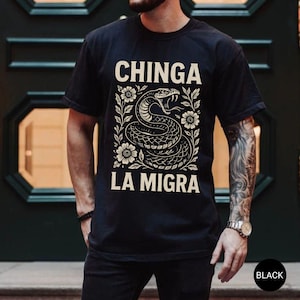 Puede incluir: Camiseta negra con un dise&ntilde;o de serpiente y el texto "CHINGA LA MIGRA" en una fuente decorativa. La serpiente est&aacute; rodeada de elementos florales. La camiseta es de cuello redondo.