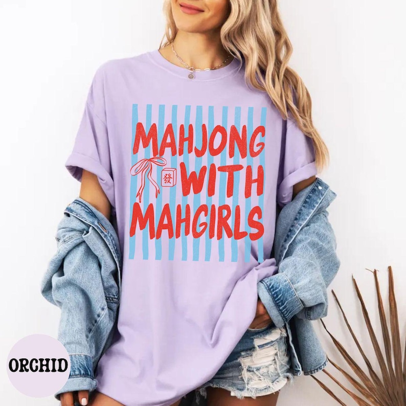 Peut inclure: Un t-shirt violet clair avec un motif vertical de rayures bleu clair et un texte rouge indiquant MAHJONG WITH MAHGIRLS, accompagn&eacute; d'un ruban rouge et d'une petite ic&ocirc;ne de tuile de mah-jong rouge avec un caract&egrave;re chinois sur le devant.