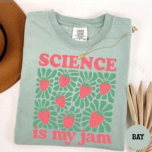 May include: A sage green short-sleeve t-shirt featuring a graphic print of pink strawberries and green leaves. The text SCIENCE is printed in pink above the strawberries, and the text is my jam is printed in pink below the strawberries.