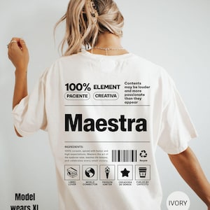 May include: An ivory t-shirt featuring a black graphic on the back. The design resembles a nutrition label with the text Maestra, 100% ELEMENT, PACIENTE, CREATIVA, and a list of ingredients. It includes a barcode, a recycle icon, and five small icons representing a book, globe, torch, star, and coffee cup with labels.