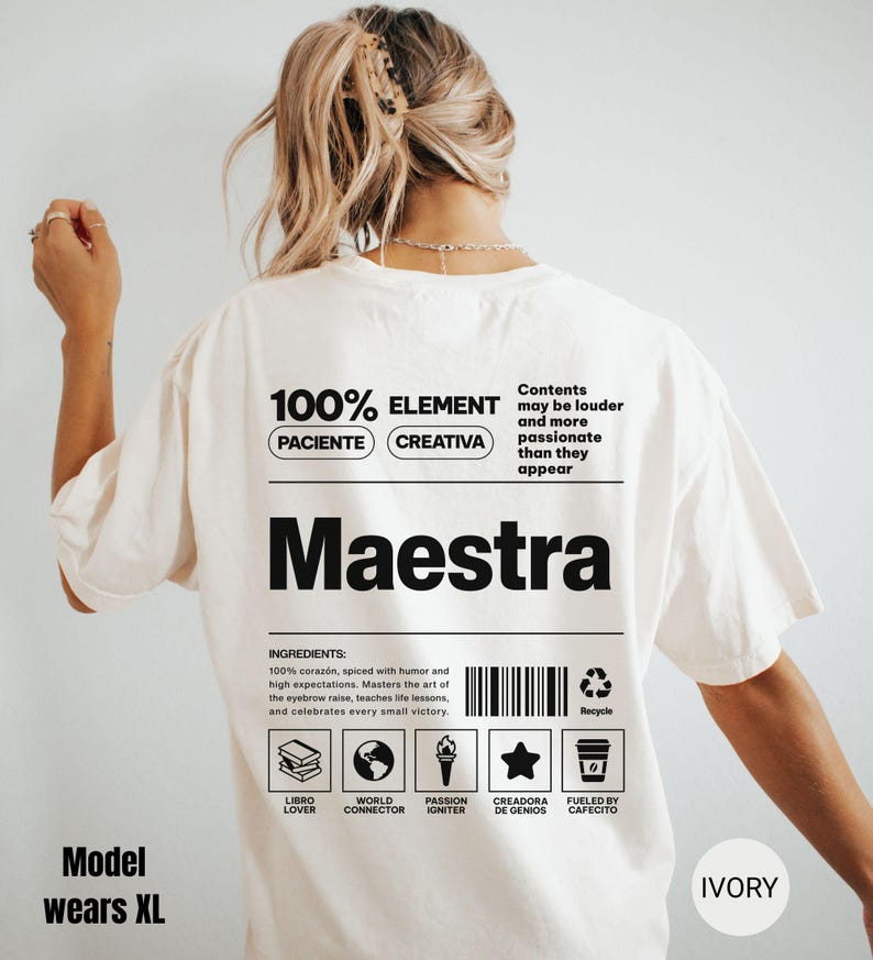 May include: An ivory t-shirt featuring a black graphic on the back. The design resembles a nutrition label with the text Maestra, 100% ELEMENT, PACIENTE, CREATIVA, and a list of ingredients. It includes a barcode, a recycle icon, and five small icons representing a book, globe, torch, star, and coffee cup with labels.