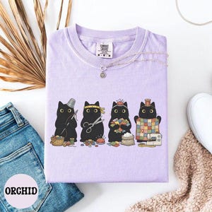 May include: A light purple t-shirt featuring a graphic of four black cats engaged in sewing activities. The cats are depicted with sewing tools including a thimble, scissors, a pincushion, and a quilt. The shirt has a crew neck and a Comfort Colors label.