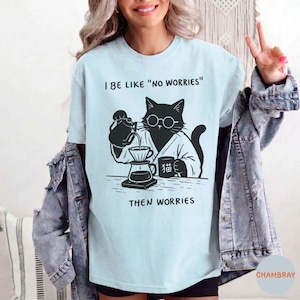 May include: A light blue t-shirt featuring a black graphic of a cat wearing round glasses, pouring coffee into a dripper, with the text I BE LIKE "NO WORRIES" and THEN WORRIES, and a mug with the Japanese character for cat printed on it.