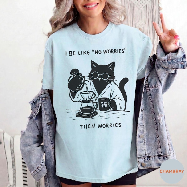 May include: A light blue t-shirt featuring a black graphic of a cat wearing round glasses, pouring coffee into a dripper, with the text I BE LIKE "NO WORRIES" and THEN WORRIES, and a mug with the Japanese character for cat printed on it.