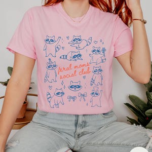 K&ouml;nnte beinhalten: Rosafarbenes T-Shirt mit einem blauen Aufdruck von Waschb&auml;ren in verschiedenen Posen und dem Text "feral moms social club". Das Design umfasst Herzen und Schleifen. Das T-Shirt wird mit blauen Jeans und wei&szlig;en Turnschuhen getragen.