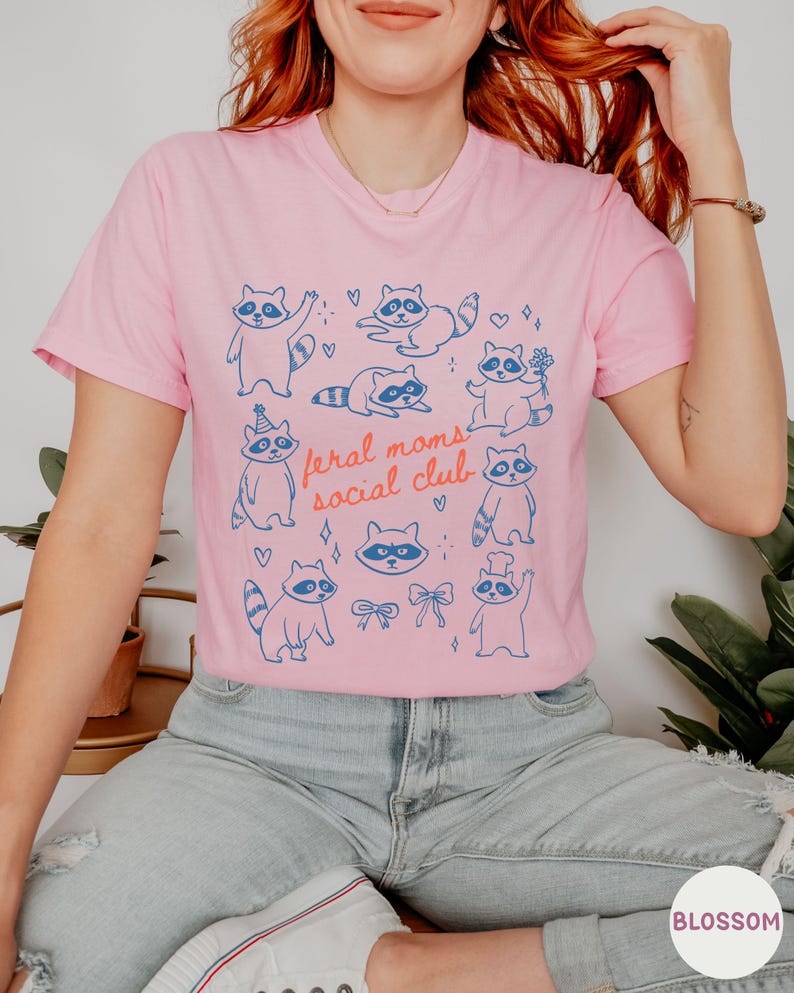 K&ouml;nnte beinhalten: Rosafarbenes T-Shirt mit einem blauen Aufdruck von Waschb&auml;ren in verschiedenen Posen und dem Text "feral moms social club". Das Design umfasst Herzen und Schleifen. Das T-Shirt wird mit blauen Jeans und wei&szlig;en Turnschuhen getragen.