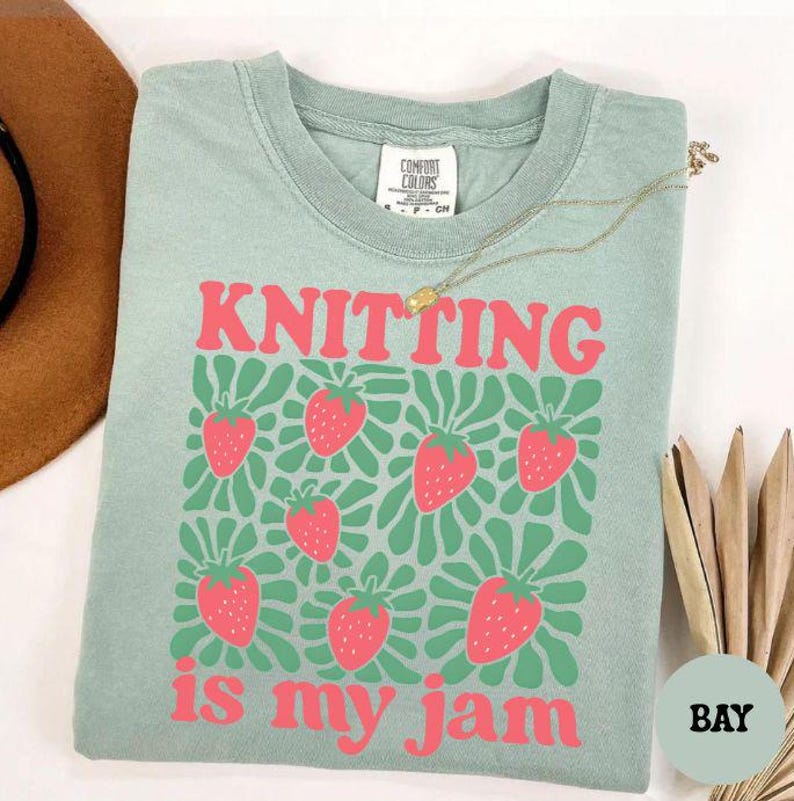 May include: A light green t-shirt with the words "KNITTING is my jam" in pink, along with a pattern of pink strawberries and green leaves. The shirt has a crew neck and short sleeves. A gold necklace and a brown hat are also visible.