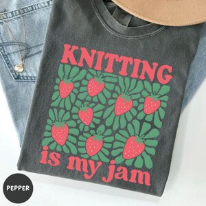 May include: Dark gray t-shirt with the text "KNITTING" in red above a graphic of strawberries and green leaves. Below the graphic, the text "is my jam" is in red. The shirt is folded on top of blue jeans.