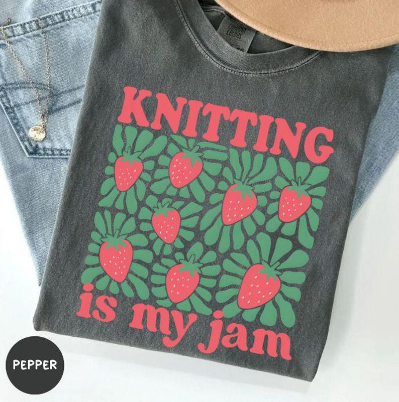 May include: Dark gray t-shirt with the text "KNITTING" in red above a graphic of strawberries and green leaves. Below the graphic, the text "is my jam" is in red. The shirt is folded on top of blue jeans.