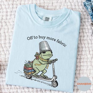 May include: Light blue t-shirt with a whimsical illustration of a frog riding a scooter, wearing a thimble hat, and carrying fabric and sewing supplies. The text "Off to buy more fabric" is printed above the frog.