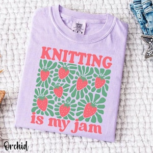 May include: Orchid-colored t-shirt with the words "KNITTING is my jam" in red. The shirt features a pattern of strawberries and green leaves. The shirt is made of a soft material and is folded.