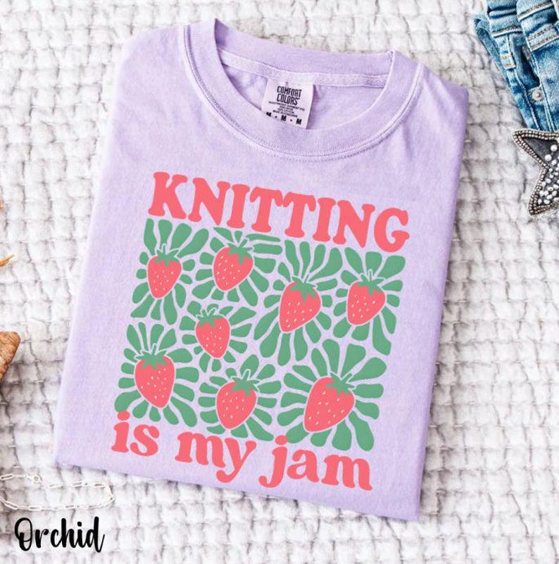 May include: Orchid-colored t-shirt with the words "KNITTING is my jam" in red. The shirt features a pattern of strawberries and green leaves. The shirt is made of a soft material and is folded.