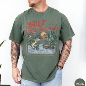 May include: Moss green t-shirt with a retro graphic. The graphic features a lake scene with a fisherman in a boat, a jumping fish, and the text "MILF Man I Love Fish" in red and orange. The shirt is a short-sleeved crew neck.