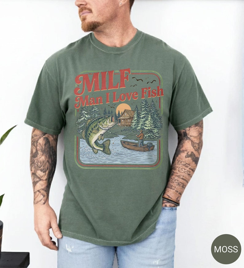 May include: Moss green t-shirt with a retro graphic. The graphic features a lake scene with a fisherman in a boat, a jumping fish, and the text "MILF Man I Love Fish" in red and orange. The shirt is a short-sleeved crew neck.