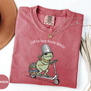 May include: Crimson-colored t-shirt with a whimsical frog graphic riding a scooter, wearing a thimble hat and carrying spools of thread. The text "Off to buy more fabric" is arched above the frog. A gold necklace and a hat are visible.