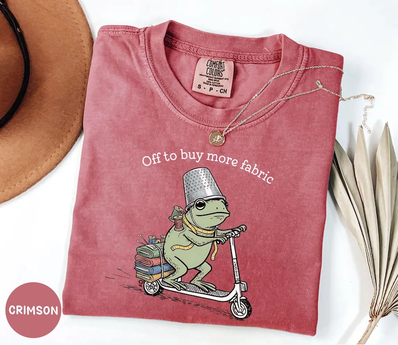 May include: Crimson-colored t-shirt with a whimsical frog graphic riding a scooter, wearing a thimble hat and carrying spools of thread. The text "Off to buy more fabric" is arched above the frog. A gold necklace and a hat are visible.